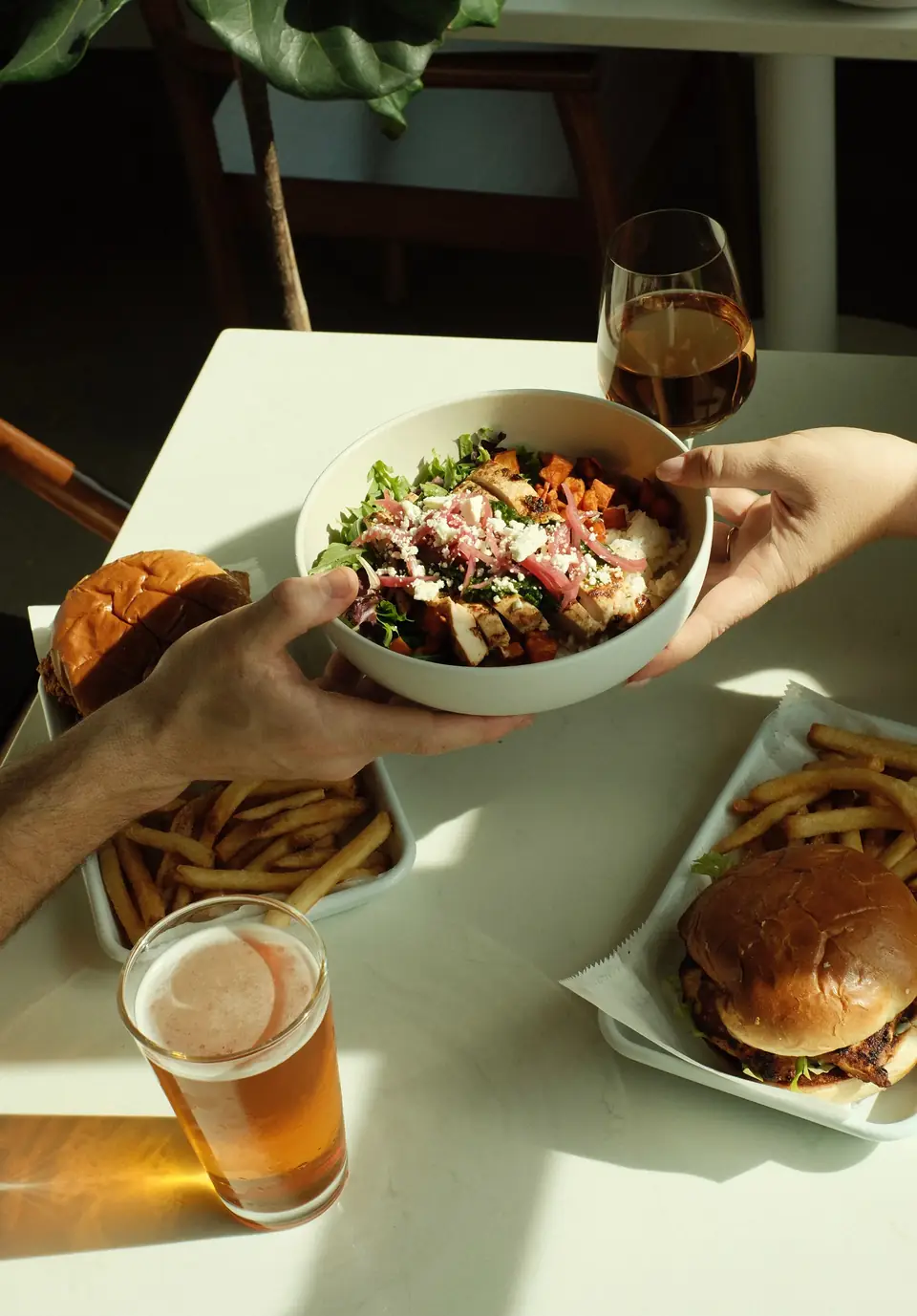 Friends passing a bowl across a sunny table of burgers, bowls and cold beer